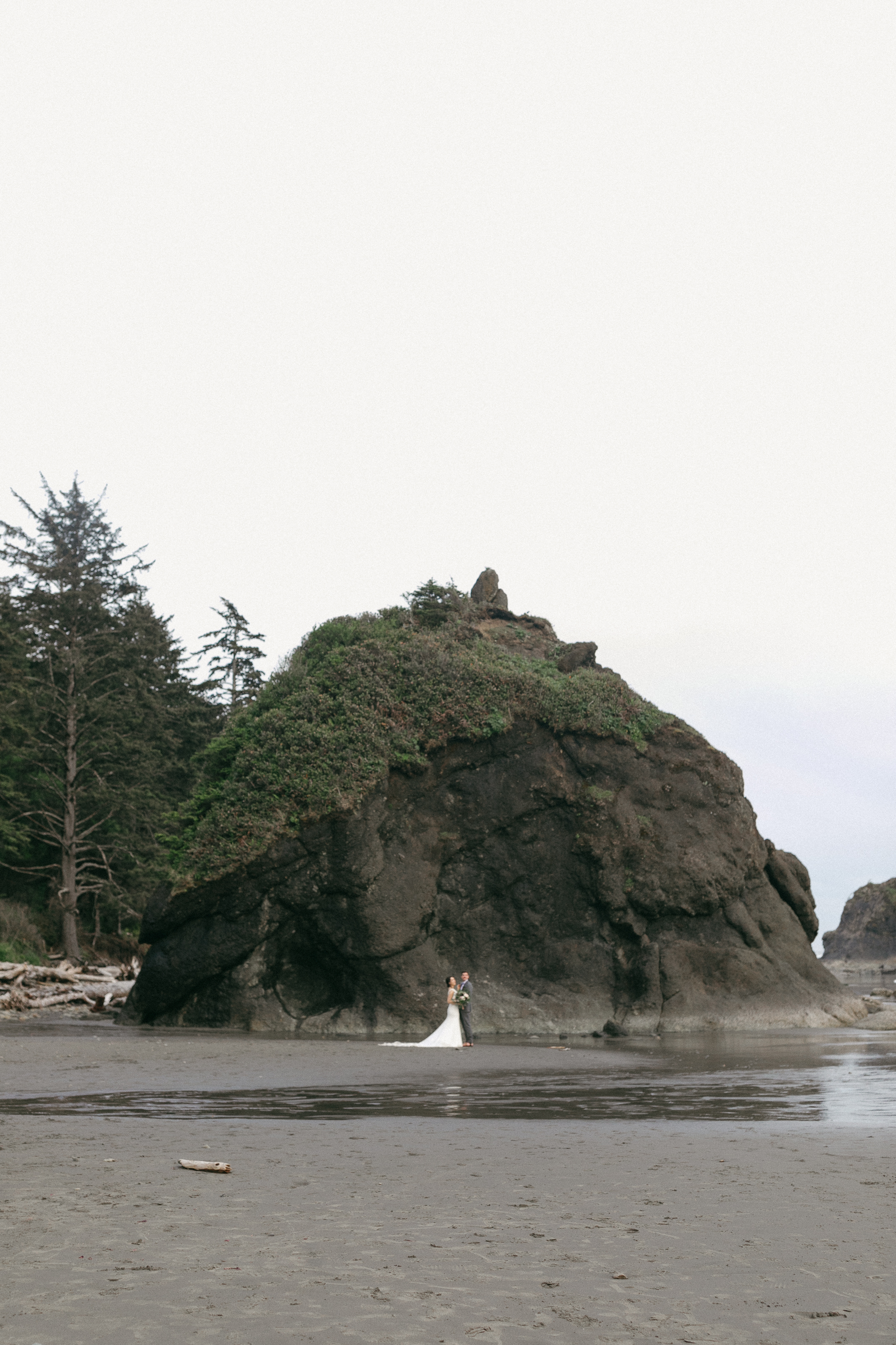 A couple stands tiny beside a towering sea stack on an empty beach while eloping in Olympic National Park, Washington